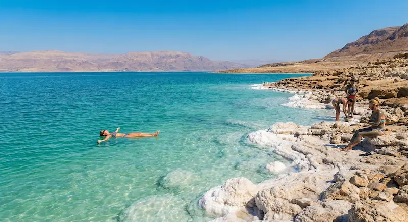Split screen comparison showing glowing bioluminescent beach at night and person floating in Dead Sea during day
